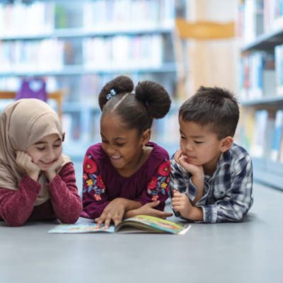 Three (3) kids who are friends are laying on the floor in between bookshelves in the library. They are reading a book together. They are all smiling and enjoying their afternoon.